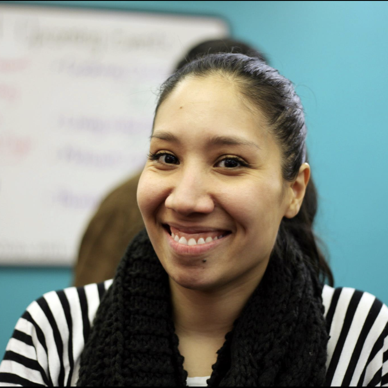 Charlotte Logan in a black-and-white striped shirt, in front of a bright blue wall with a whiteboard on it.
