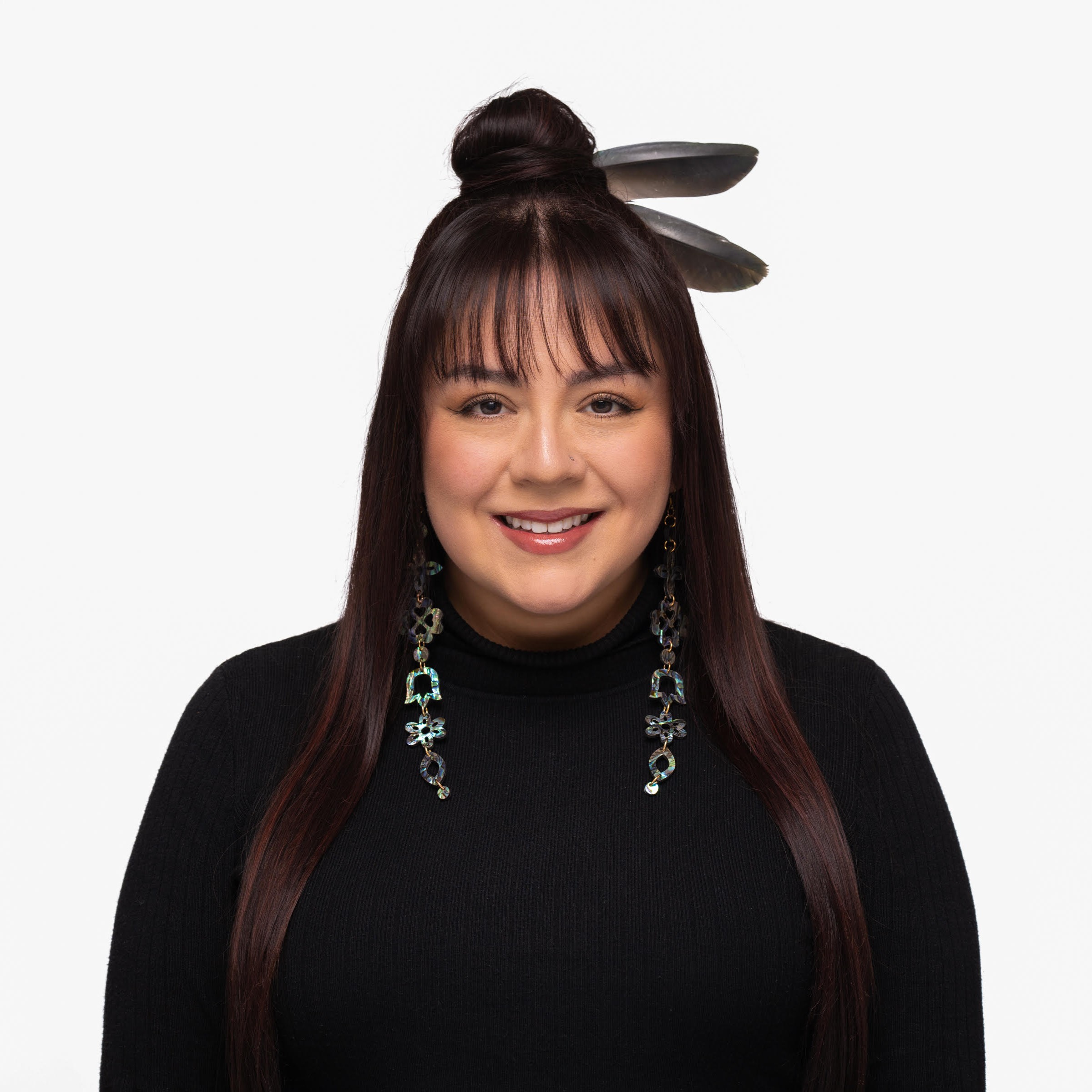Christina Thomas dressed in a black shirt with two feathers in her hair, standing against a white background.