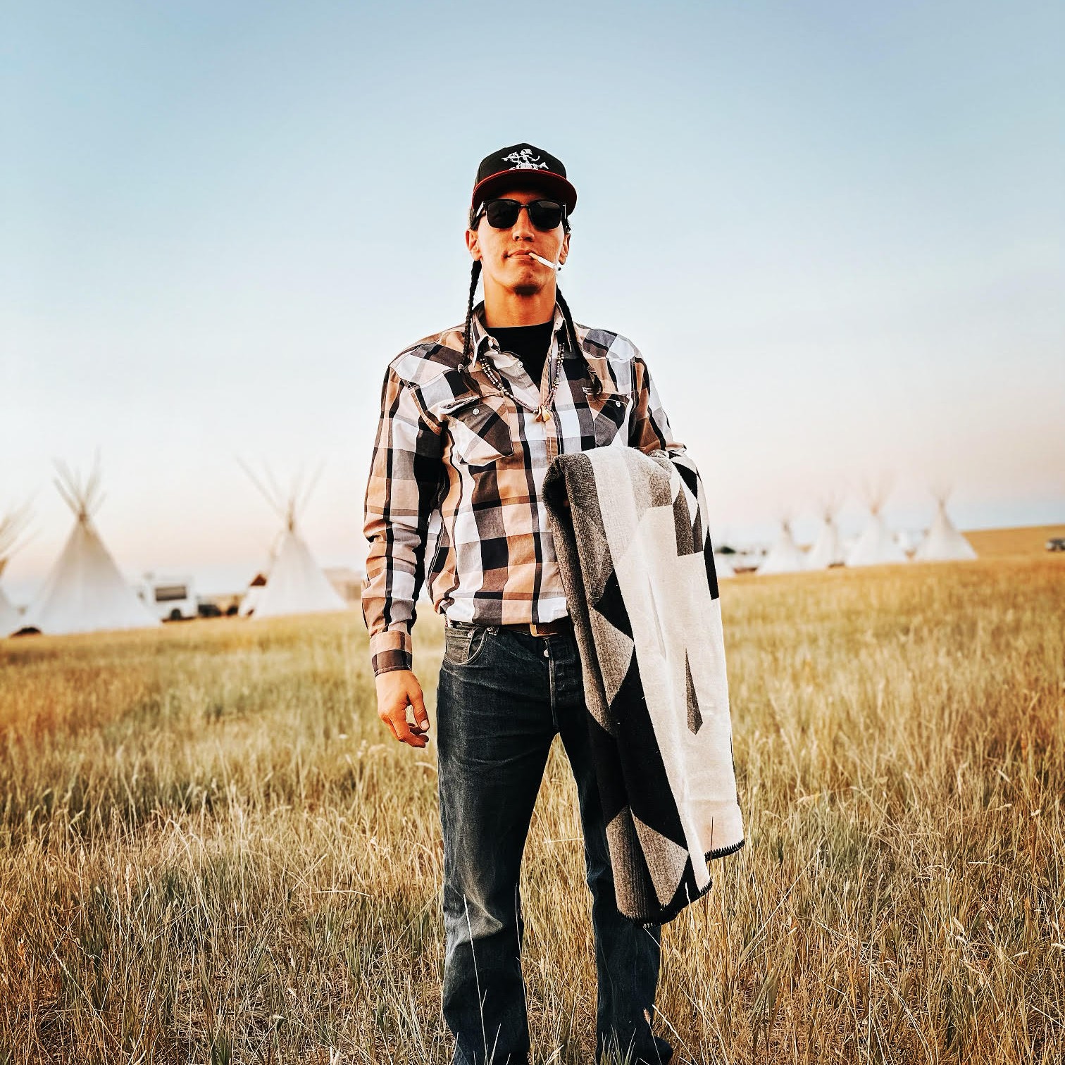 Jesse DesRosier in jeans, flannel shirt, cap and sunglasses, smoking, standing in grassland with tipis in the background holding a blanket.