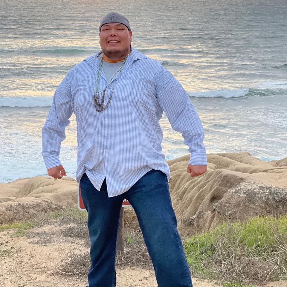 Mosiah Bluecloud in blue jeans and a blue long-sleeve shirt standing on sand dunes in front of the ocean.
