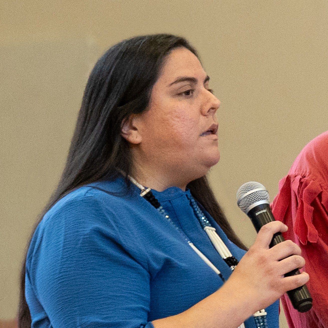 Elisabeth Mora in a blue shirt and necklace holding a microphone and speaking