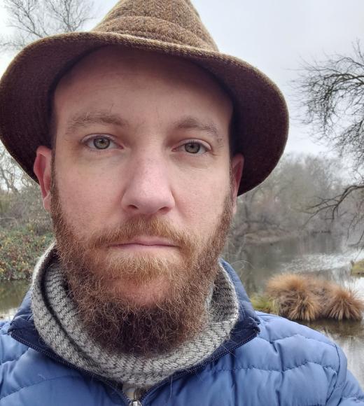 Lewis Lawyer headshot with tweed hat, scarf and puffer jacket, with Putah Creek in the background.
