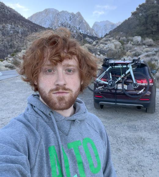 Duncan Tyler in a grey and green sweatshirt, outside in the mountains, car with bike in the background