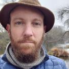 Lewis Lawyer headshot with tweed hat, scarf and puffer jacket, with Putah Creek in the background.