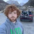 Duncan Tyler in a grey and green sweatshirt, outside in the mountains, car with bike in the background